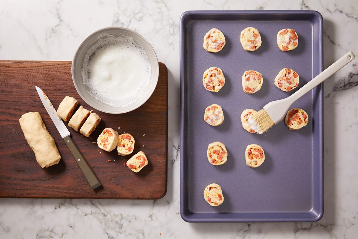 Overhead horizontal image of sliced pepperoni pinwheels arranged on a baking sheet, with visible layers of dough, cheese, and pepperoni. A pastry brush and egg wash are styled nearby.