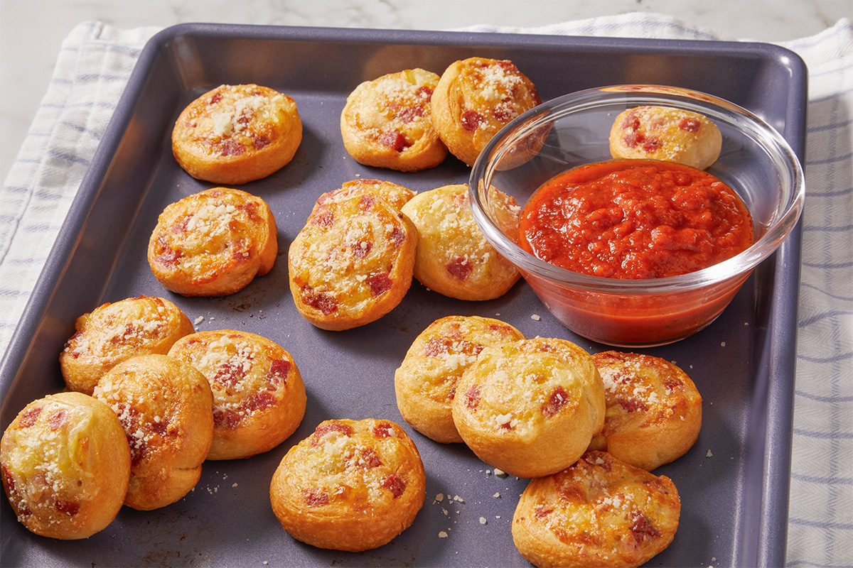 Overhead horizontal image of baked pepperoni pinwheels sprinkled with grated Parmesan cheese and served alongside marinara sauce. The presentation highlights texture, browning, and melted cheese.