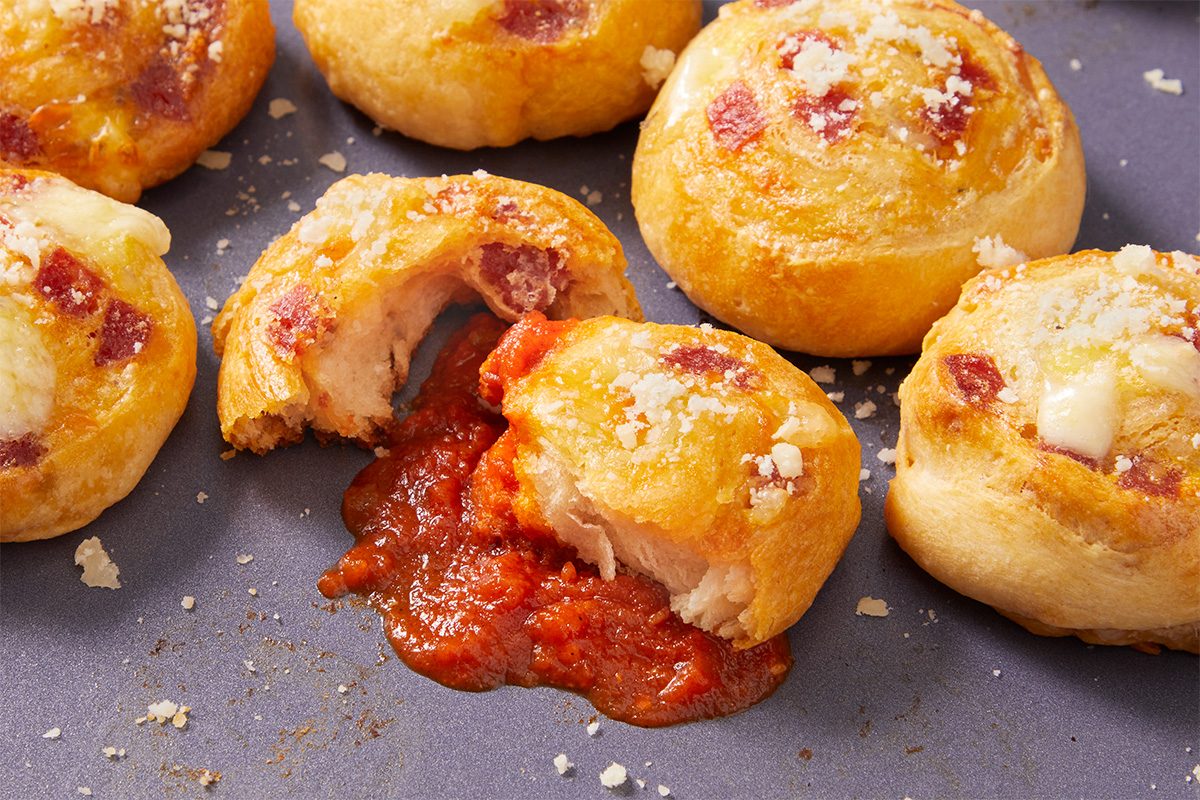 Overhead horizontal close-up image of a pepperoni pinwheel broken open to reveal melted mozzarella and pepperoni, with marinara sauce spilling from the center for visual impact.