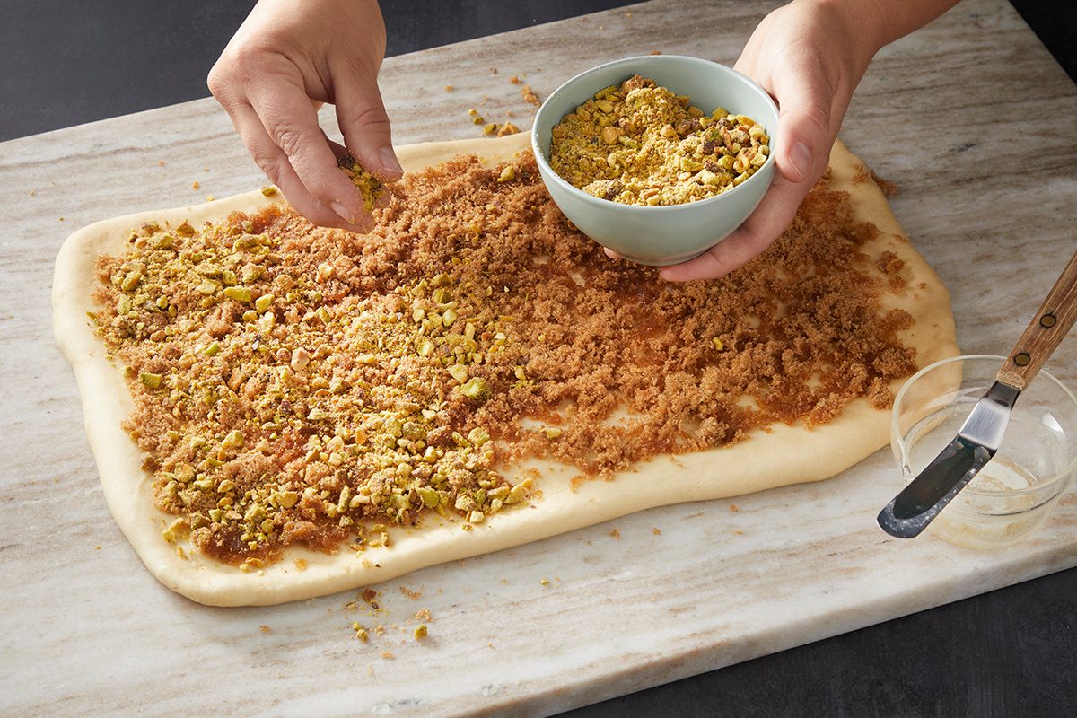 A person sprinkles chopped nuts and brown sugar over a rectangular sheet of dough on a marble surface, preparing it for baking.