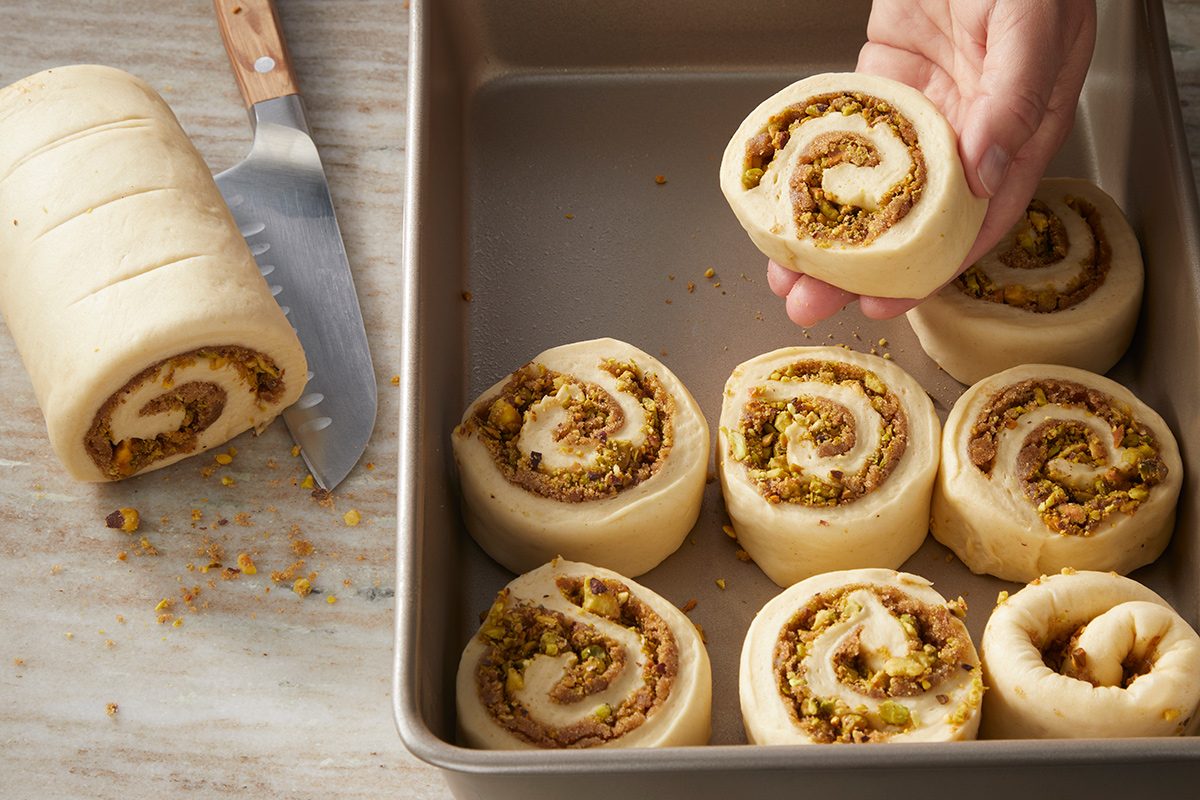 A hand places a raw, unbaked nut-filled pastry swirl into a baking pan with several similar pastries. A knife and a log of sliced dough are nearby on a light countertop.