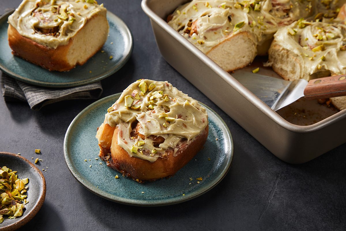 A close-up of frosted cinnamon rolls topped with chopped pistachios, served on green plates and in a baking pan, with a small bowl of pistachios on the side.