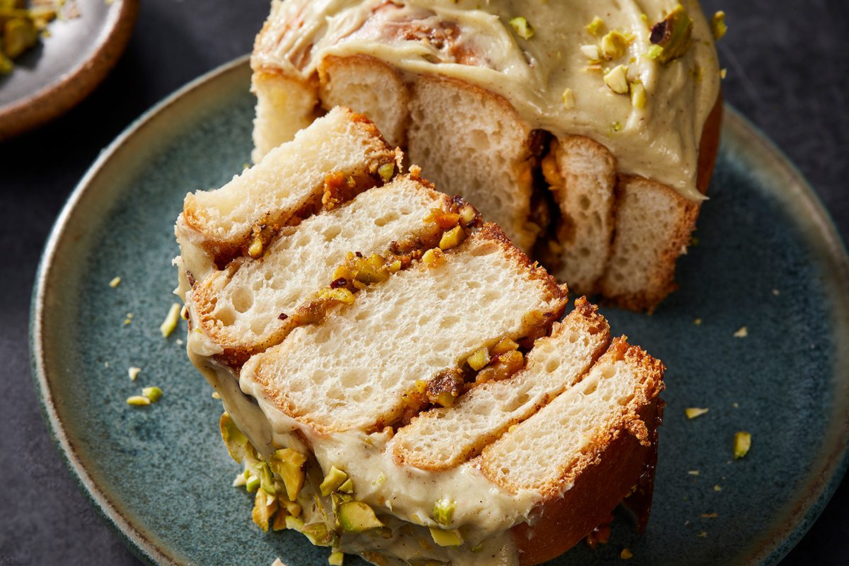 A close-up of a sliced loaf cake with creamy frosting and chopped pistachios on top, served on a blue plate. The cake shows visible layers with a nutty filling inside.