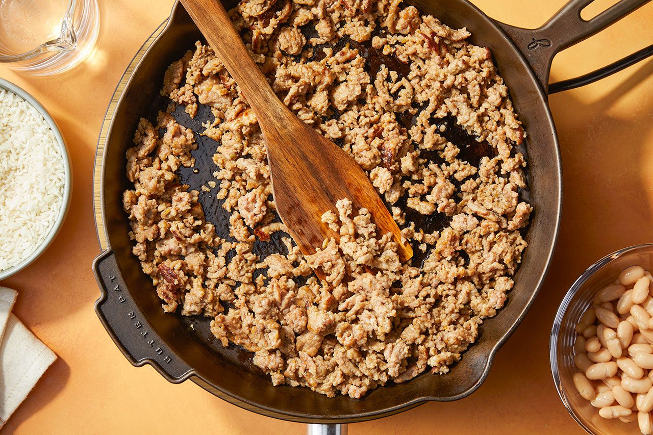 Overhead horizontal image of browned bulk Italian sausage cooking in a cast-iron skillet with a wooden spoon resting in it. To the left is a bowl of uncooked instant rice, and to the right is a bowl of cannellini beans, with a measuring cup of water at the top.