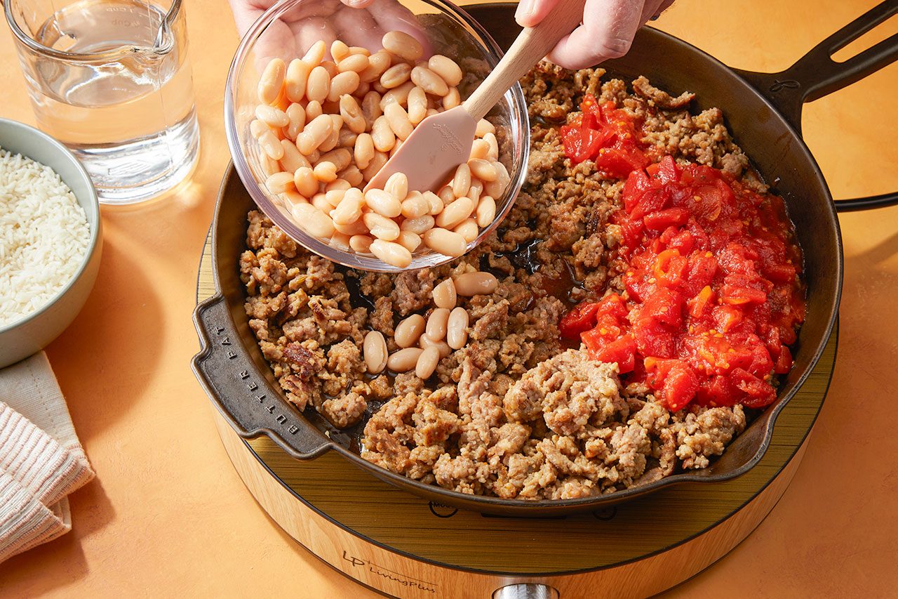 Overhead horizontal image of a hand adding cannellini beans from a glass bowl into a black skillet. The skillet already contains browned Italian sausage and diced tomatoes. In the background, a bowl of uncooked instant rice and a measuring cup with water are visible.