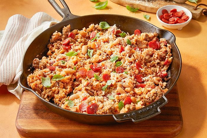 Eye Level horizontal image of the finished Meat Lover's Pizza Rice Skillet in a black cast iron pan. The skillet is filled with a hearty mixture of cooked rice, Italian sausage, cannellini beans, diced tomatoes, and sliced mini pepperoni, garnished with grated Parmesan cheese and fresh basil leaves. A small white bowl of extra sliced mini pepperoni is visible in the background.