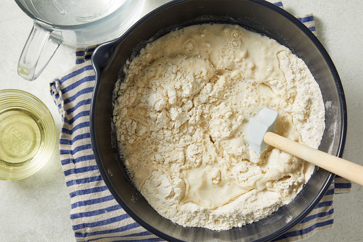Overhead horizontal image of ingredients being mixed in a black bowl with a rubber spatula. Flour is being combined with a liquid to create a dough, as part of a recipe step.