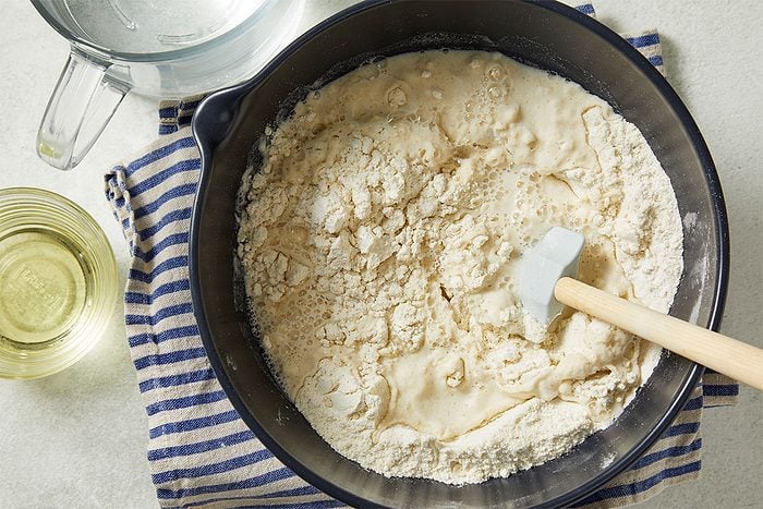 Overhead horizontal image of ingredients being mixed in a black bowl with a rubber spatula. Flour is being combined with a liquid to create a dough, as part of a recipe step.