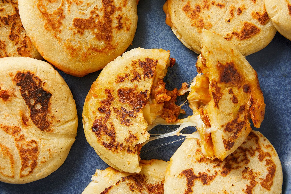 Overhead horizontal image of arepas. One of the arepas is torn open, showing ground meat and stringy melted cheese inside.