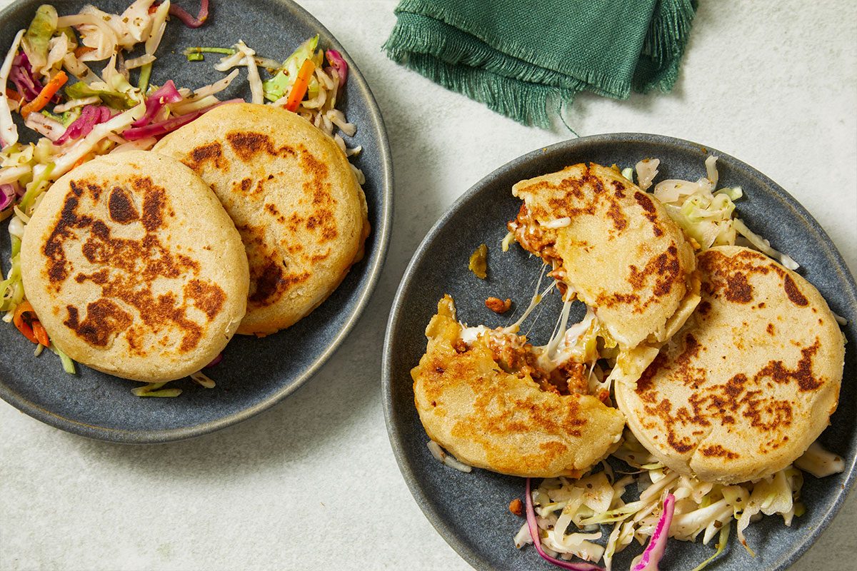 Overhead horizontal image of two plates of pupusas and curtido. One pupusa is broken open revealing melted cheese inside, as well as visible cabbage and carrots in the slaw.