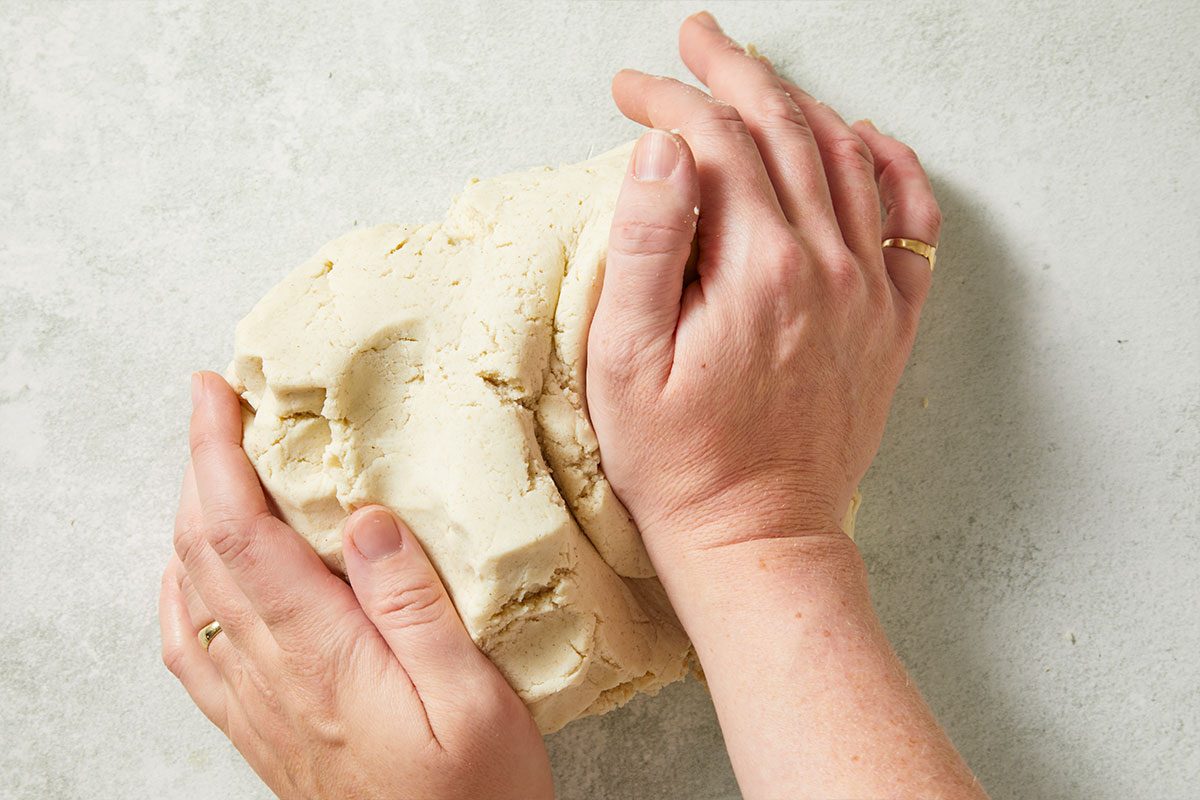 Overhead horizontal image of dough being kneaded by two hands on a countertop. The dough is pale in color. The hands have gold rings on their ring fingers.