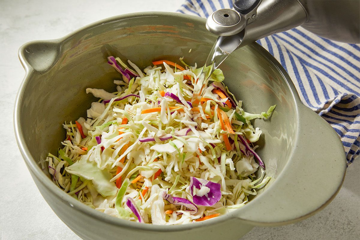 Eye Level horizontal image of coleslaw mix in a gray bowl. Water is being poured from a metal container into the bowl of cabbage, carrots, and purple cabbage.