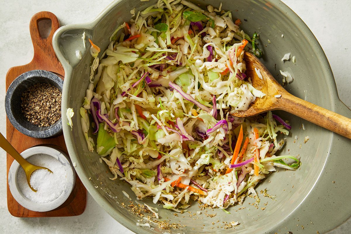 Overhead horizontal image of coleslaw being mixed in a green ceramic bowl with a wooden spoon. Shredded cabbage, carrots, and purple cabbage are visible in the bowl, along with salt and pepper nearby.