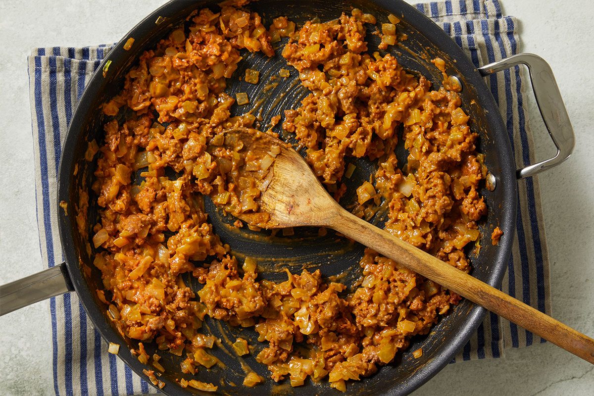 Overhead horizontal image of ground meat and onions in a skillet, being stirred with a wooden spoon. A blue and white striped towel is under the corner of the pan.