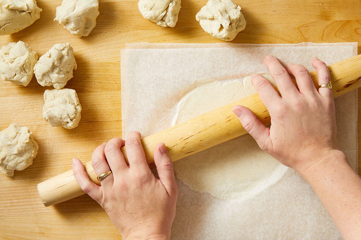 Overhead horizontal image of dough being rolled with a wooden rolling pin on parchment paper. Several small clumps of dough sit nearby on a wooden surface.