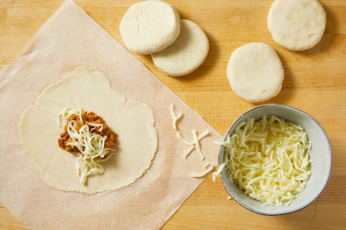 Overhead horizontal image of the process of filling a homemade tortilla with meat filling and shredded cheese. The tortilla is sitting on parchment paper. A bowl of shredded cheese sits to the right of the tortilla and a few filled and closed tortillas sit in the top right corner.