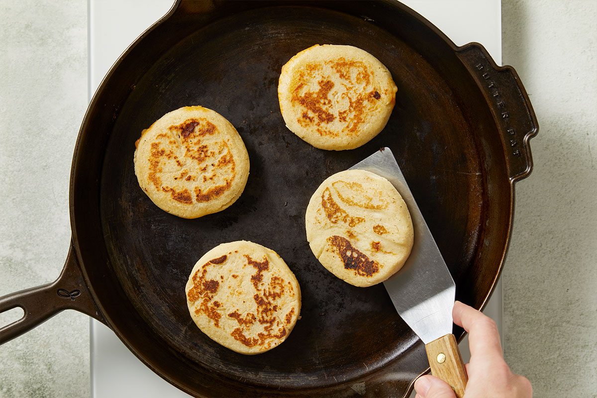 Overhead horizontal image of four arepas being flipped in a cast iron skillet. A hand holds a metal spatula with a wooden handle, ready to flip one of the arepas. The arepas are golden brown with slightly crisp exteriors.