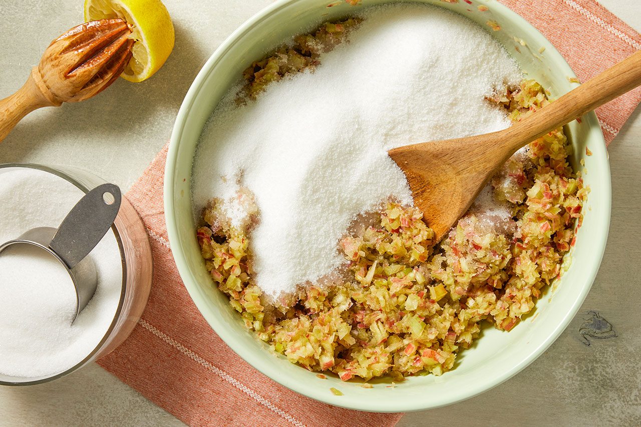 Overhead shot of a large mixing bowl containing a finely chopped fruit mixture topped with granulated sugar, as a wooden spoon stirs the ingredients together. A lemon juicer, measuring cup of sugar, and a kitchen towel are arranged nearby on a light surface.
