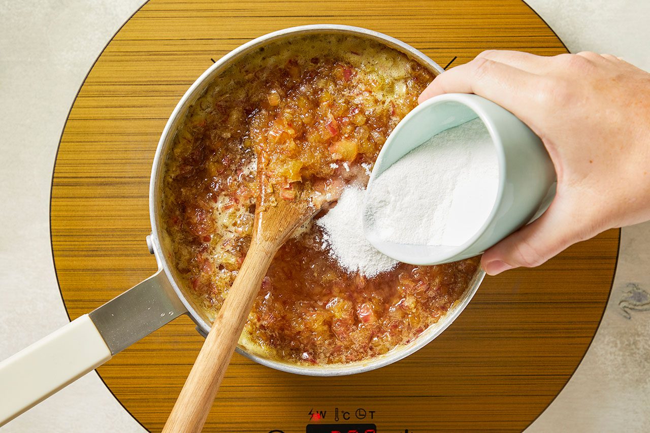 Overhead shot of a fruit mixture cooking in a saucepan on an induction cooktop as granulated sugar is poured in from a small bowl. A wooden spoon stirs the mixture while the cooktop display is visible beneath the pan.