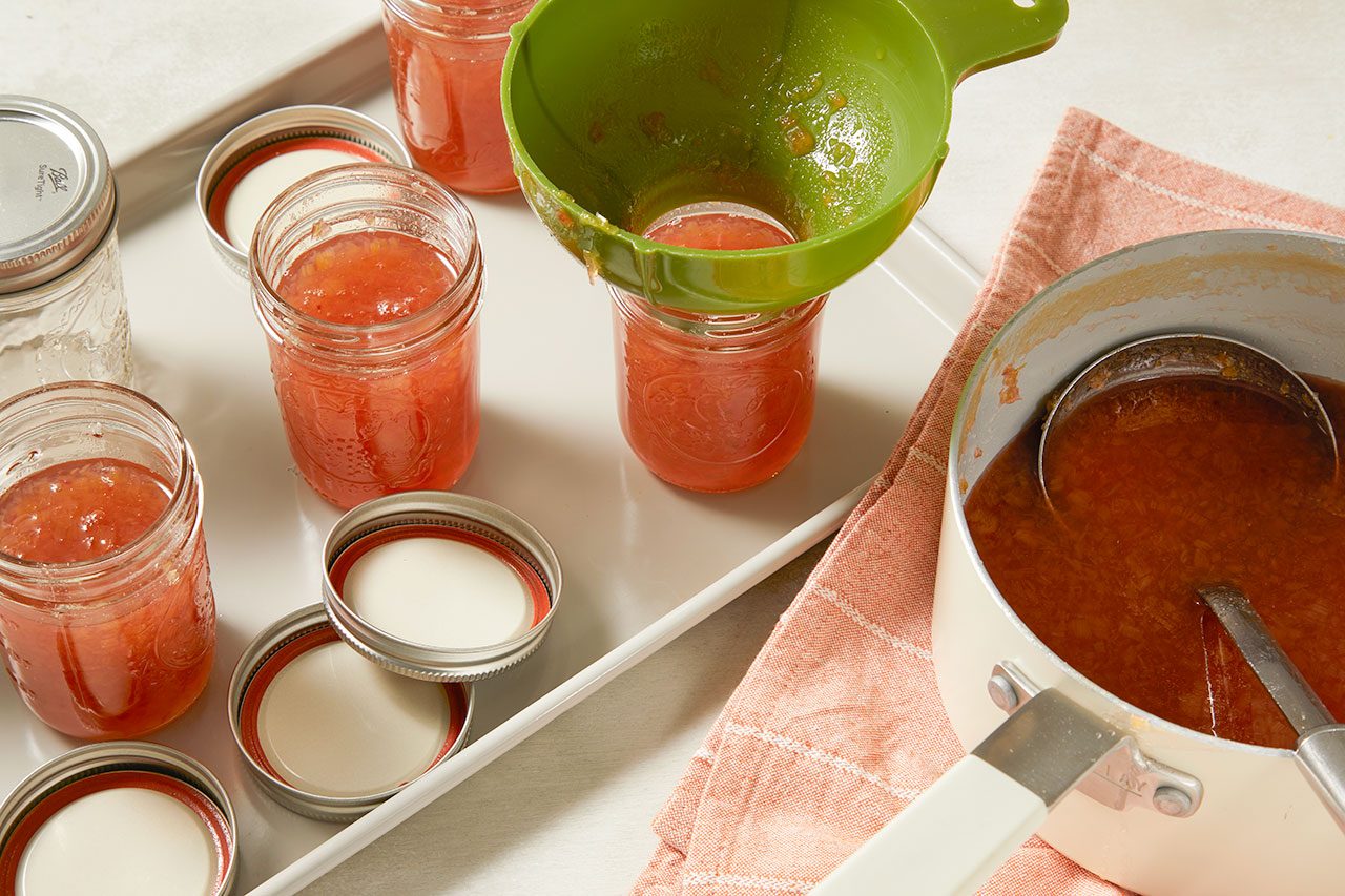 3/4 angle view shot of fruit preserves being poured through a green canning funnel into glass jars, with additional filled jars, canning lids, and a saucepan of hot preserves arranged nearby on a light countertop.