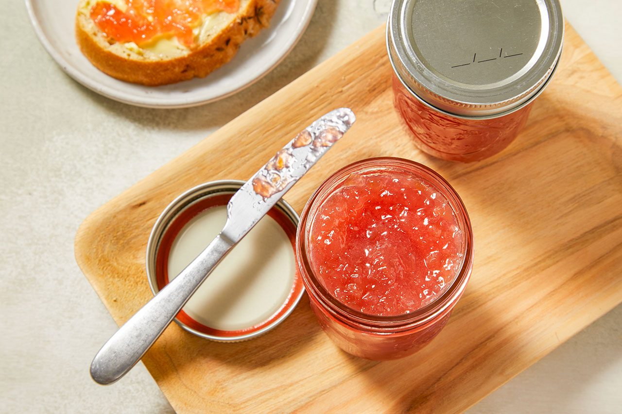 Overhead shot of Rhubarb Freezer Jam in glass mason jars, highlighting its vibrant pink color and chunky texture. One open jar sits beside a canning lid and butter knife on a wooden cutting board, with a slice of toast spread with jam visible in the background.
