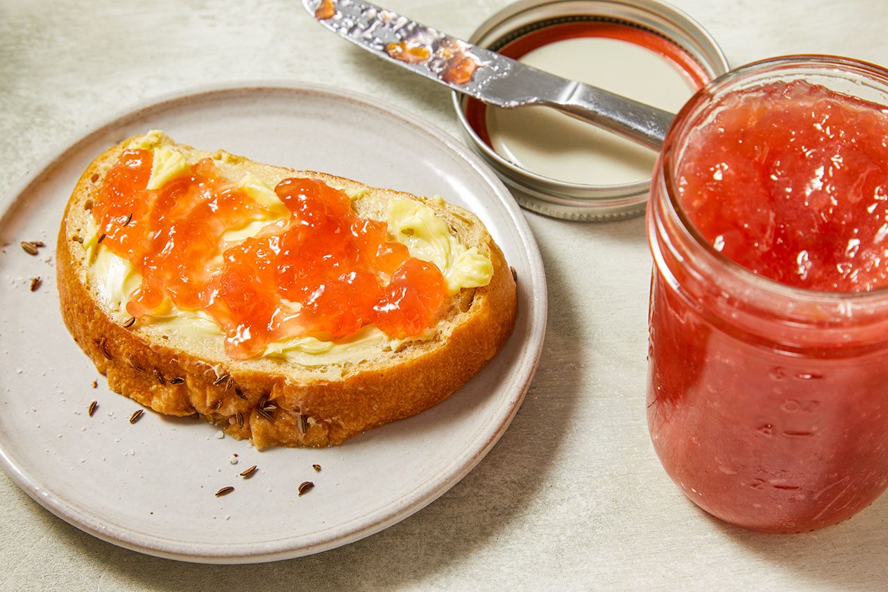 Close-up shot of Rhubarb Freezer Jam spread over a slice of buttered toast, with an open glass mason jar of vibrant pink jam, a butter knife, and a canning lid arranged nearby on a light countertop.