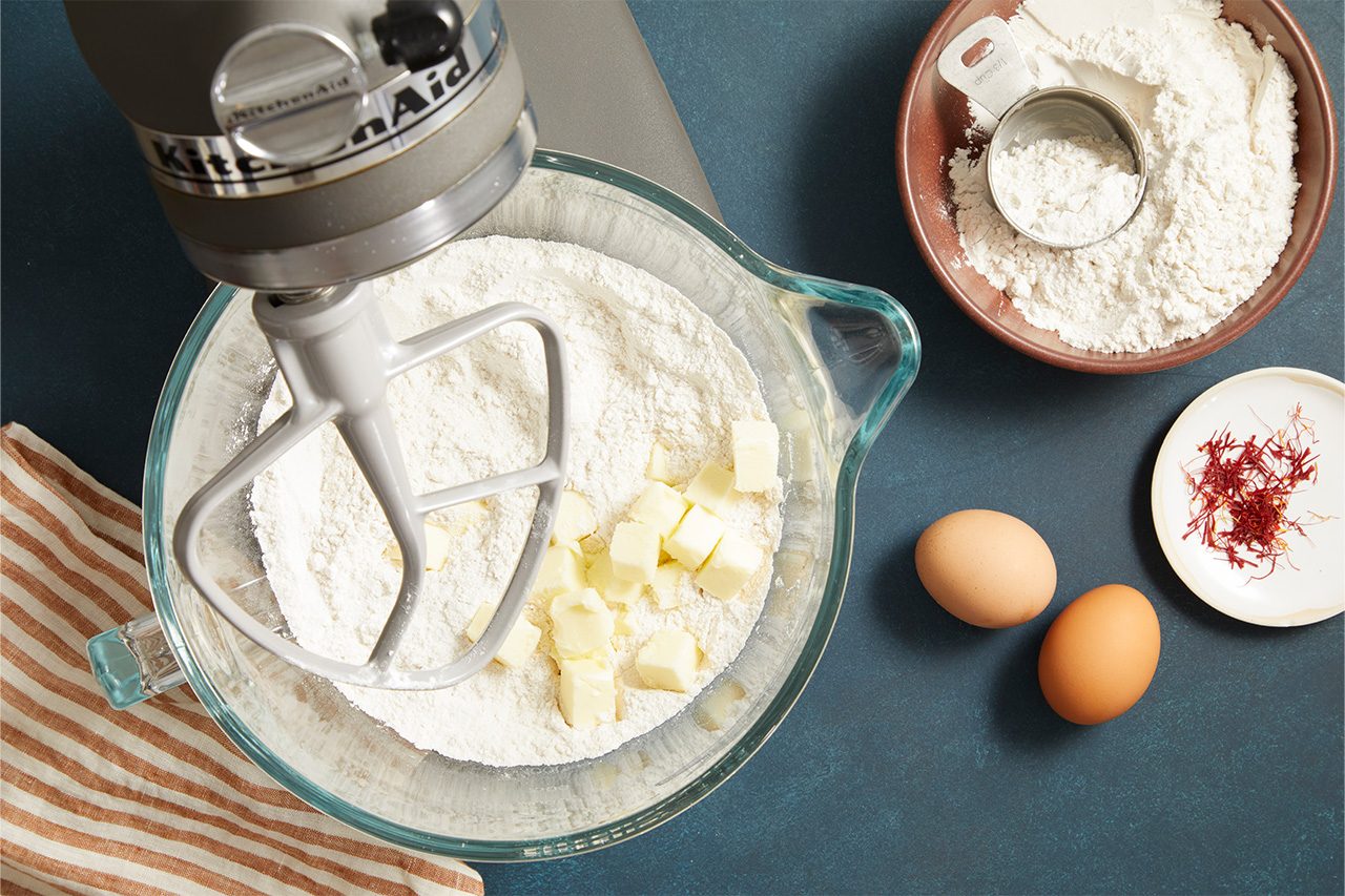 Overhead shot of a stand mixer with a glass mixing bowl containing flour and cubed butter beneath a paddle attachment. Nearby are a bowl of flour with a measuring scoop, two eggs, a small dish of red spice, and a striped kitchen towel arranged on a dark countertop;
