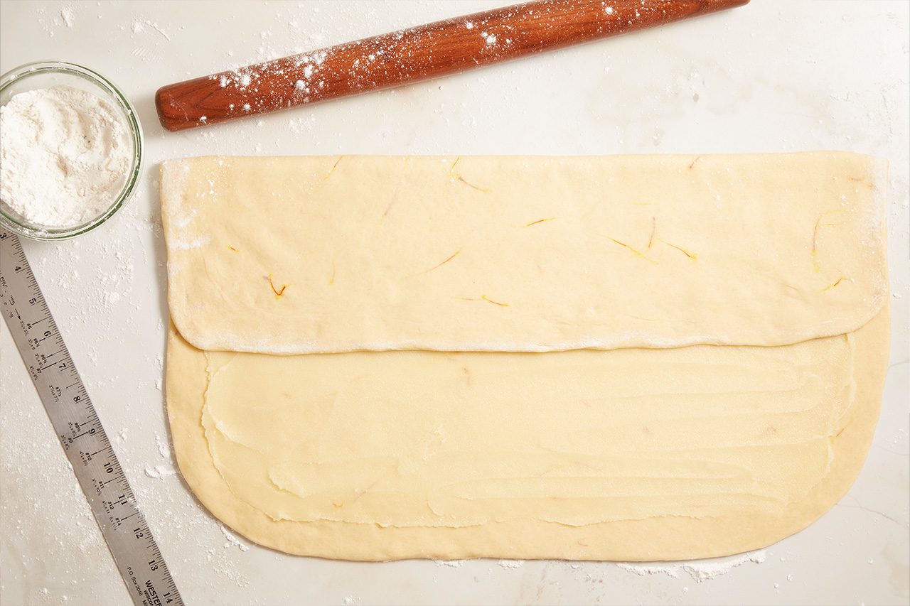 Overhead shot of a large sheet of rolled-out dough folded over itself on a lightly floured work surface, with a rolling pin, ruler, and a bowl of flour arranged nearby;