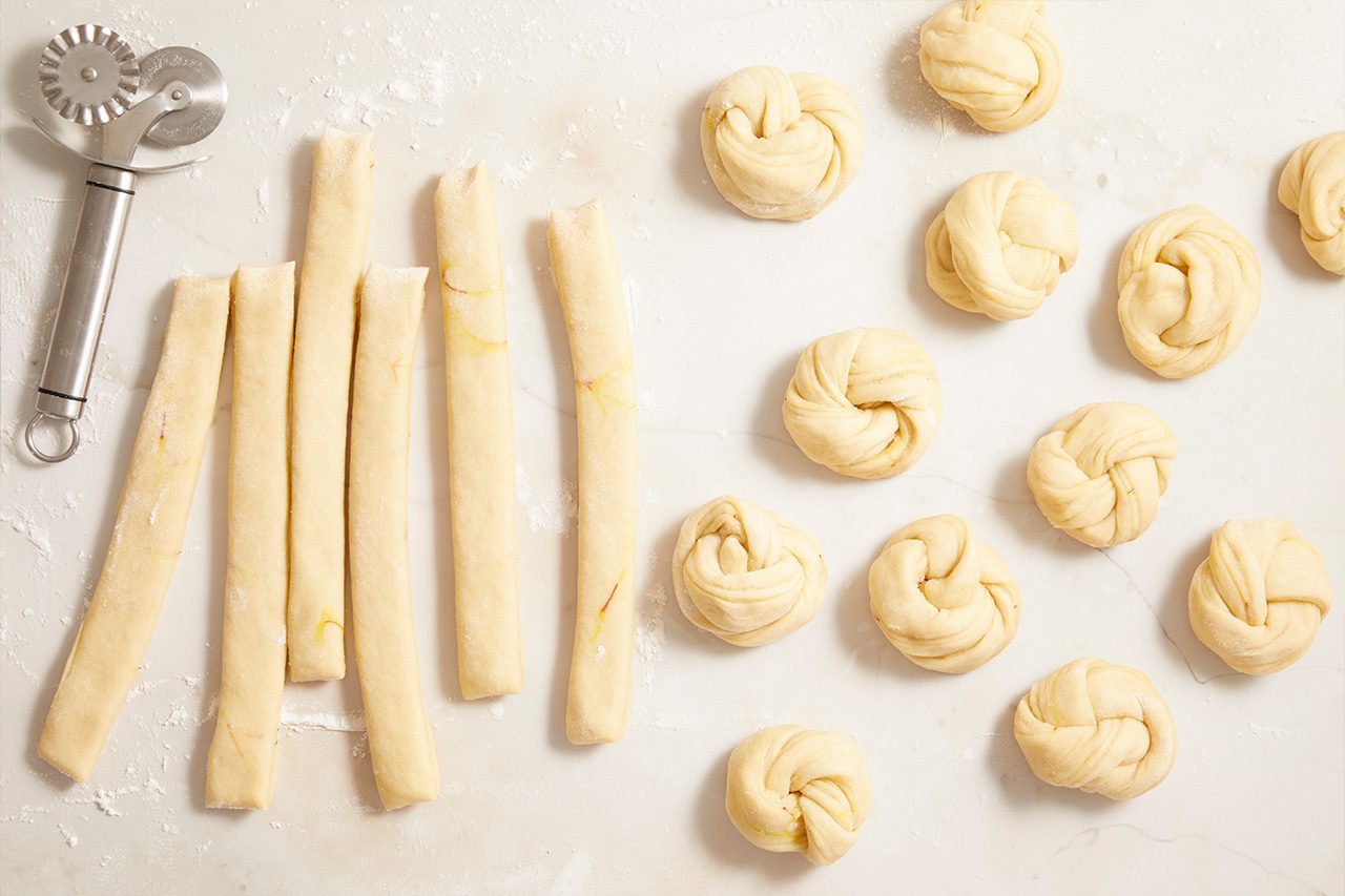 Overhead shot of strips of dough and neatly twisted dough knots arranged on a lightly floured work surface, with a pastry wheel cutter positioned in the upper left corner, illustrating the dough-shaping process;
