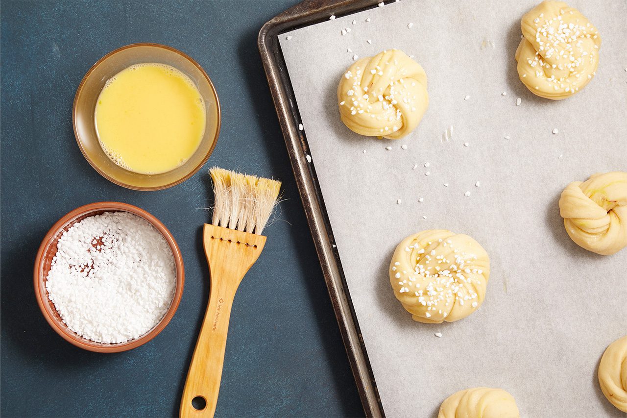 Overhead shot of unbaked twisted dough knots arranged on a parchment-lined baking sheet, sprinkled with coarse sugar. Nearby are a bowl of melted butter, a bowl of granulated sugar, and a pastry brush for coating the dough before baking;