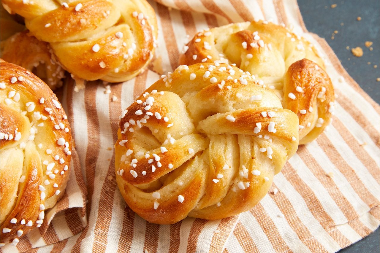 Close-up shot of a freshly baked saffron bun with a glossy golden-brown crust, twisted knot shape, and pearl sugar topping, arranged on a striped kitchen towel with additional saffron buns softly blurred in the background;