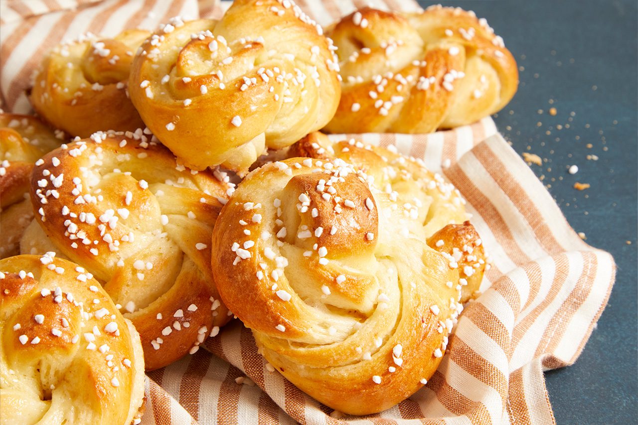 3/4 angle view shot of freshly baked saffron buns with a glossy golden-brown crust, twisted knot shape, and pearl sugar topping, arranged on a striped kitchen towel, highlighting their soft texture and rich color;