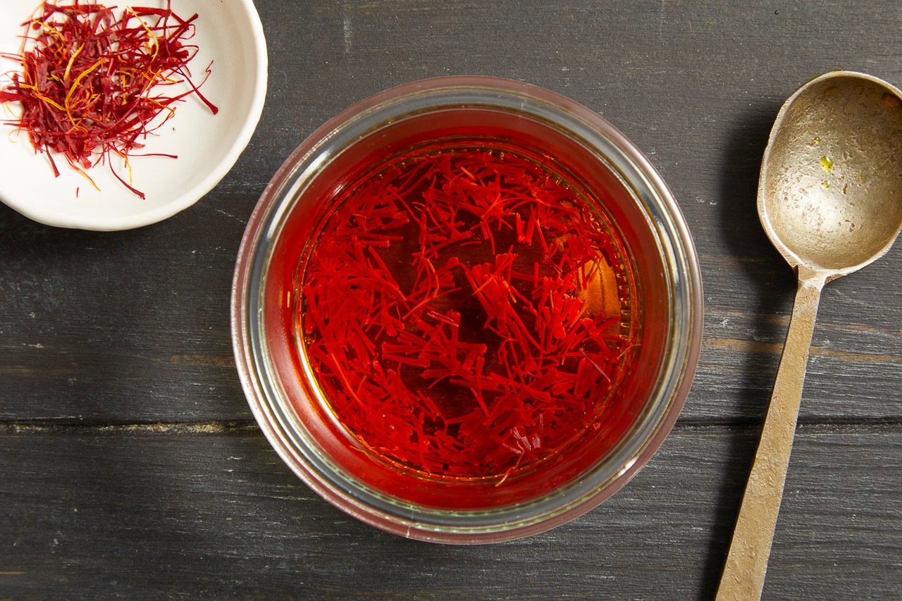 Overhead shot of a small glass bowl filled with saffron threads floating on the surface. A small dish of dried saffron threads and a vintage spoon are arranged nearby on a dark wooden surface