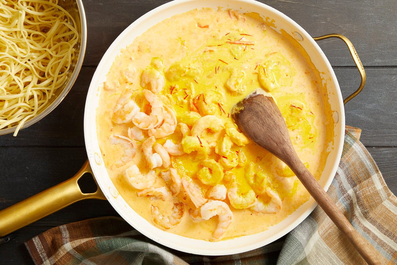 Overhead shot of shrimp simmering in a creamy saffron sauce in a white skillet, stirred with a wooden spoon
