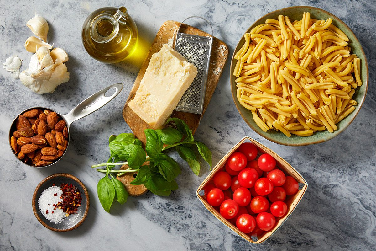 Overhead horizontal image of various fresh ingredients laid out on a marble surface. The arrangement includes garlic, a cruet of olive oil, a block of Parmesan cheese with a grater, and a bowl of pasta. Also visible are a measuring cup full of almonds, fresh basil, cherry tomatoes, and a dish with salt and red pepper flakes.
