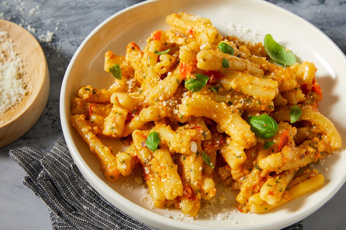 Overhead horizontal image of a white bowl filled with pasta tossed in a creamy, reddish-orange sauce. The pasta is garnished with fresh basil leaves and a generous sprinkle of grated cheese, with more grated cheese visible on a wooden dish beside the bowl.