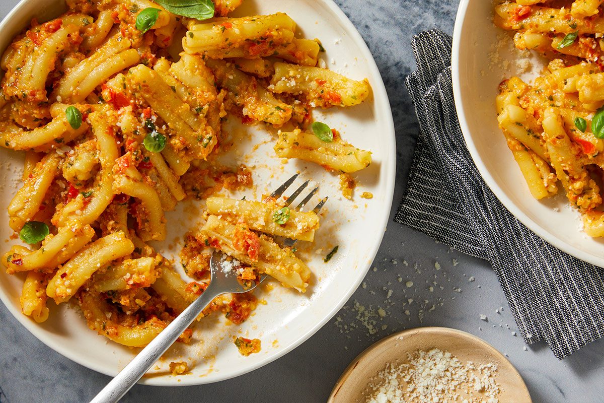 Overhead horizontal image of a white plate with short pasta tossed in a red sauce, garnished with fresh basil leaves and sprinkled grated cheese. A fork rests in the partially eaten pasta, with an additional bowl of grated cheese and a striped napkin visible on the grey surface.