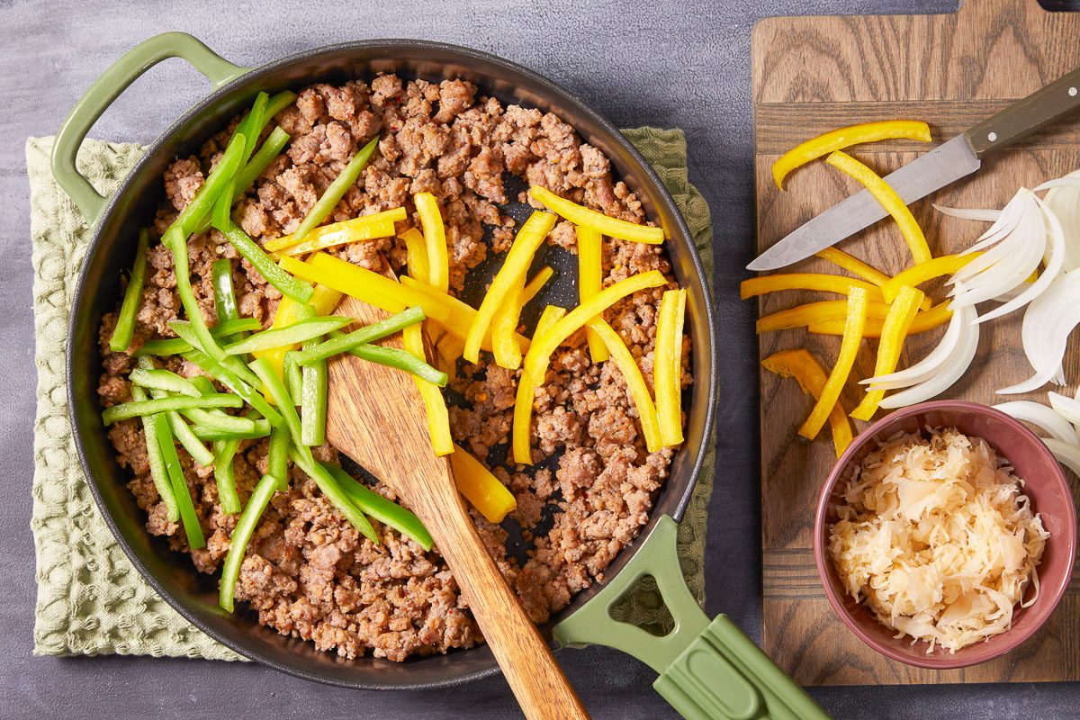 sliced bell peppers added to sausage in a skillet next to chopping board with sliced onions and bell peppers
