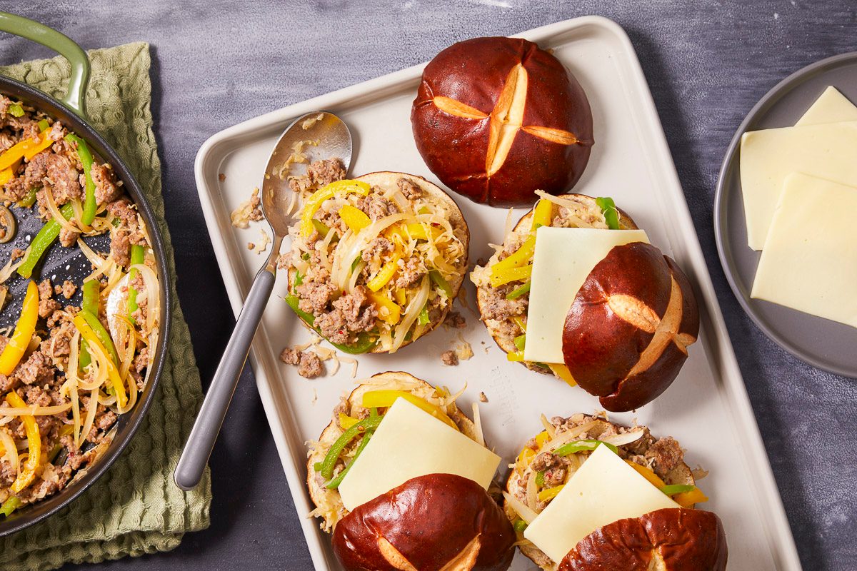 Overhead image of pretzel buns topped with a mixture of spicy pork sausage, sauerkraut, green and yellow peppers, and onions and provolone cheese. More of the meat mixture is in a pan on the side
