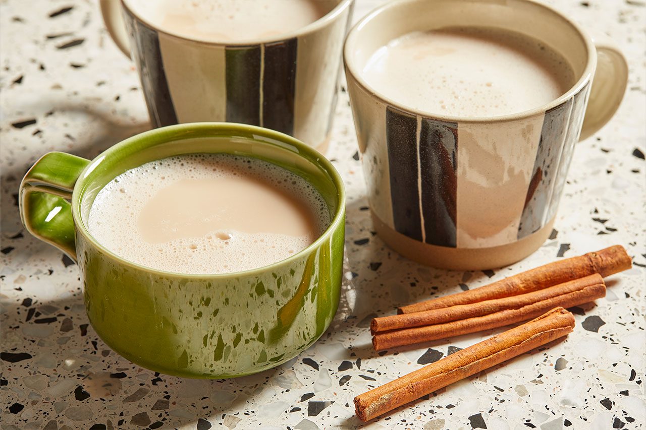 Eye Level horizontal image of three mugs filled with creamy chai tea, accompanied by three cinnamon sticks on a speckled countertop. The mugs are in shades of green and striped black and beige.