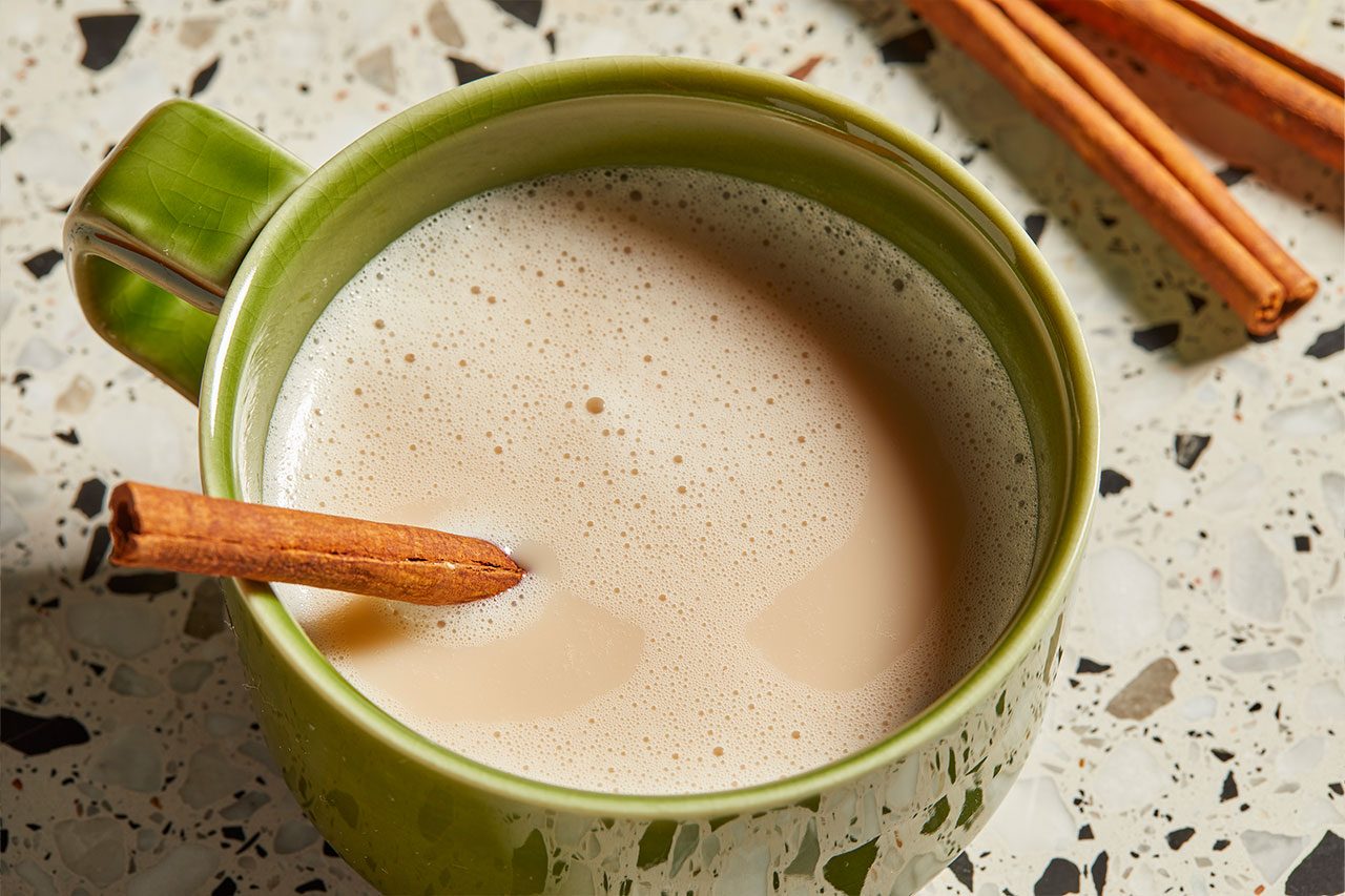 Eye level horizontal image of a green ceramic mug filled with frothed chai tea. A cinnamon stick rests partially submerged in the warm beverage, with additional cinnamon sticks visible on the speckled countertop in the background.