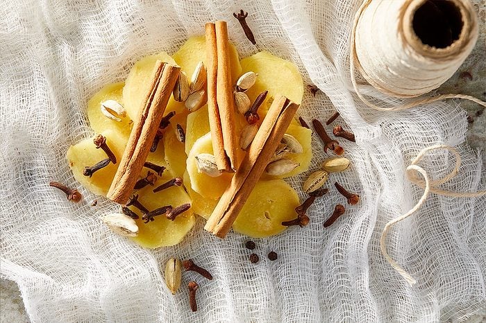Overhead horizontal image of sliced fresh ginger, cinnamon sticks, whole cloves, cardamom pods, and whole peppercorns arranged on a white cheesecloth. A spool of kitchen twine is visible in the upper right corner, with some twine unspooled onto the cheesecloth. These raw, dry ingredients are ready for making chai tea.