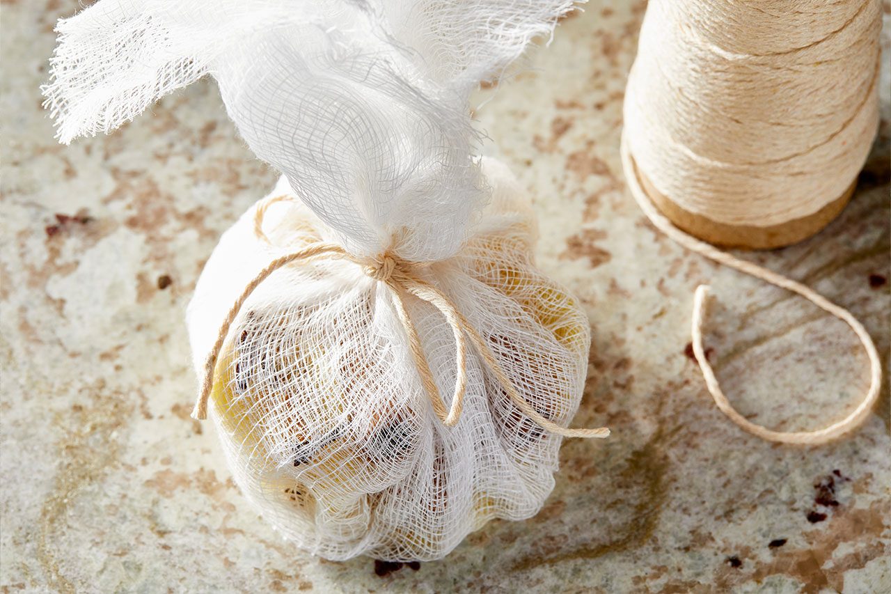 Eye level horizontal image of a bundle of spices tied in cheesecloth, next to a spool of twine. Sliced gingerroot, cinnamon sticks, whole cloves, cardamom pods, and whole peppercorns are visible through the cheesecloth.