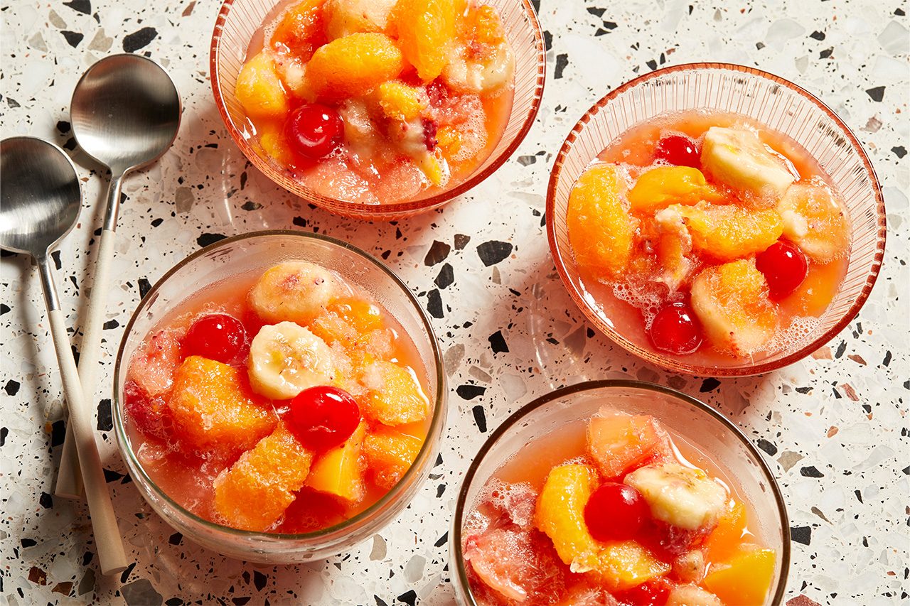 Overhead horizontal image of four clear glass bowls filled with a colorful frozen fruit slush. Each serving showcases a vibrant mix of sliced bananas, peach slices, pineapple chunks, mandarin orange segments, and bright red maraschino cherries suspended in a frosty, pinkish-red base.