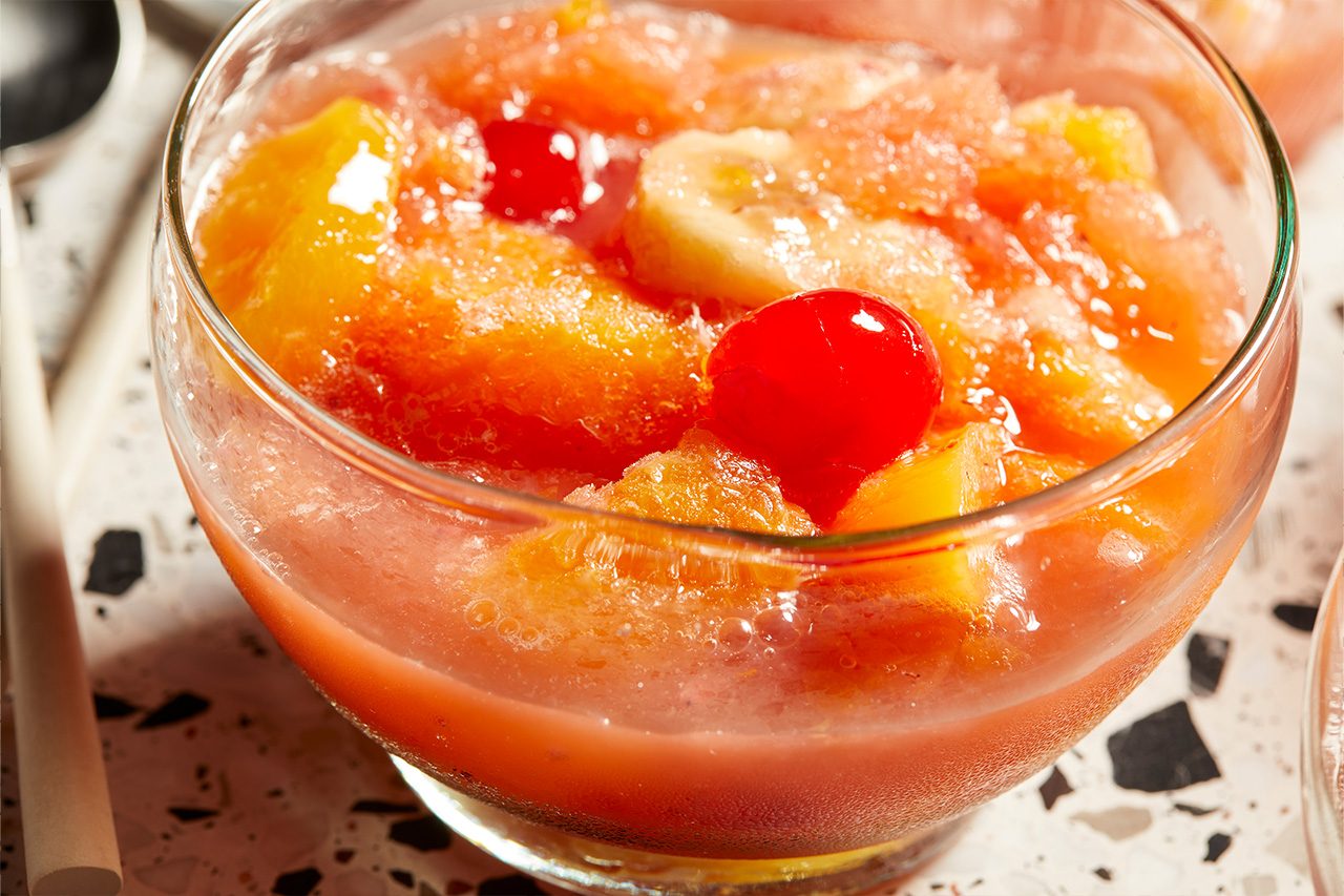 Eye Level horizontal image of a clear glass bowl filled with a colorful Frozen Fruit Slush. The slush features vibrant red maraschino cherries, slices of banana, and pieces of peach, pineapple, and mandarin orange, all suspended in a refreshing, icy mixture.