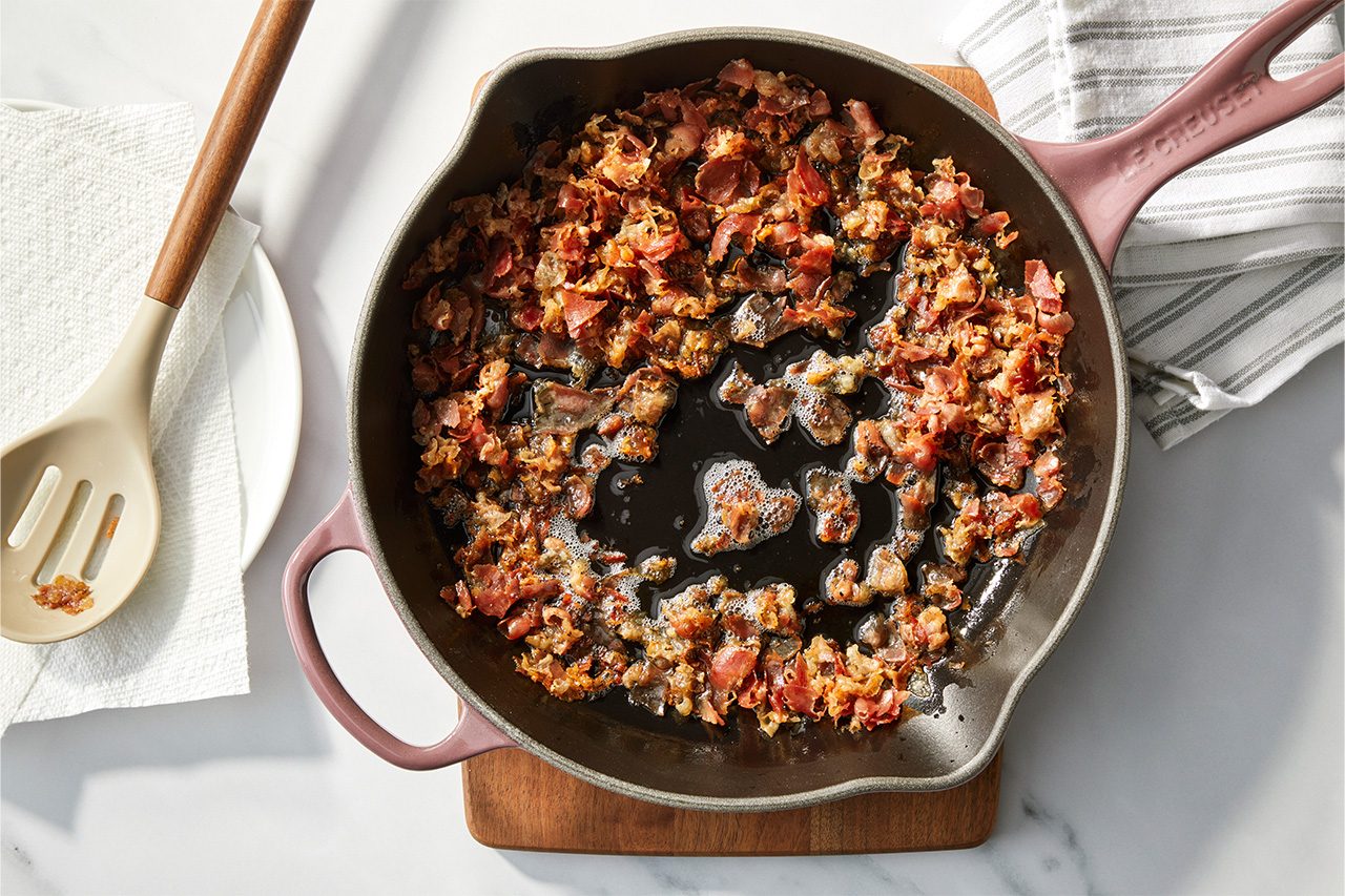 Overhead horizontal image of a purple cast iron skillet filled with crispy, cooked bacon bits and rendered fat. A beige slotted spoon with some bacon pieces rests on a paper towel and white plate to the left, while a striped kitchen towel is visible to the right. | [Original] RMS 118417 Italian Steak Bruschetta; How to; Step 1; Cooking pancetta in small skillet; Horizontal