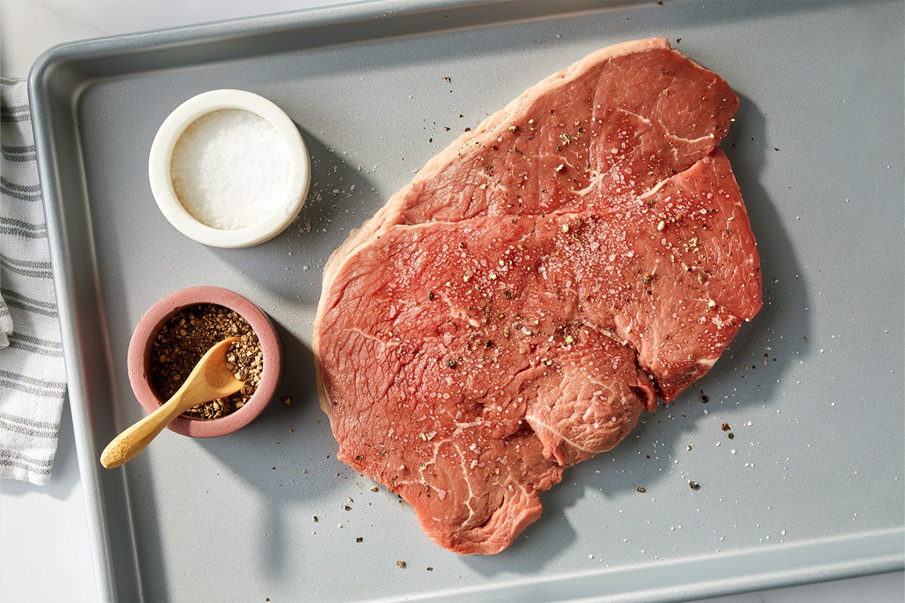 Overhead horizontal image of a raw steak seasoned with salt and pepper, lying on a gray baking sheet. A small white bowl of salt and a reddish-brown bowl of pepper with a wooden spoon are placed next to the steak, ready for seasoning. | [Original] RMS 118417 Italian Steak Bruschetta; How to; Step 2; Sprinkling steak on sheet pan with salt & pepper; Horizontal