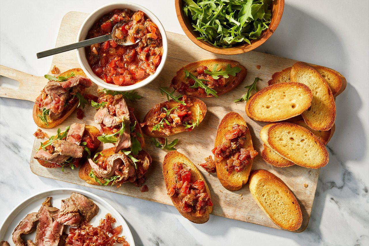 Overhead horizontal image of a wooden cutting board laden with various components for steak crostini. Toasted baguette slices are topped with either a vibrant tomato and onion mixture or thinly sliced steak with fresh arugula. A white bowl holds additional tomato mixture, while a wooden bowl contains more arugula, completing this step in the recipe. | [Original] RMS 118417 Italian Steak Bruschetta; How to; Step 3; Process style shot; French bread partially sliced on cutting board; some slices on baking sheet