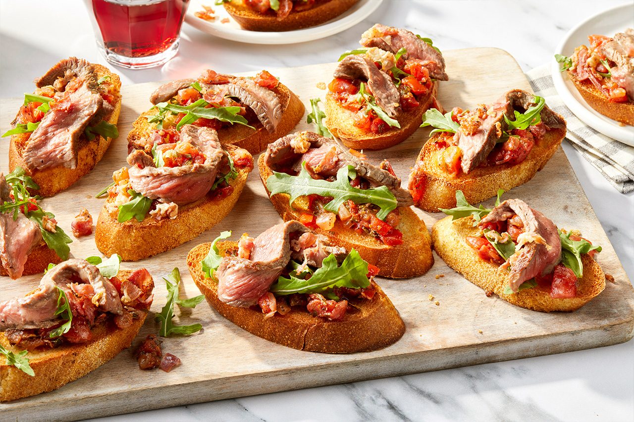 Eye Level horizontal image of multiple steak bruschetta on a wooden cutting board. Each toasted bread slice is topped with a tomato mixture, fresh arugula, and thinly sliced steak, with small pieces of bacon sprinkled on top. A glass with a red beverage is visible in the background. | [Original] RMS 118417 Italian Steak Bruschetta; Horizontal AP; Pull