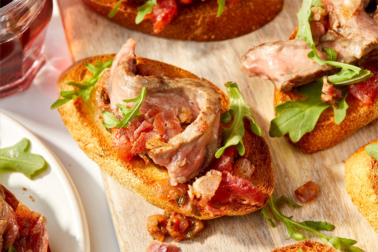 Eye level horizontal image of several crostini appetizers arranged on a wooden board. Each piece of toasted bread is topped with a rich red relish, fresh green arugula, and a slice of medium-rare beef. | [Original] RMS 118417 Italian Steak Bruschetta; How to; Horizontal Push